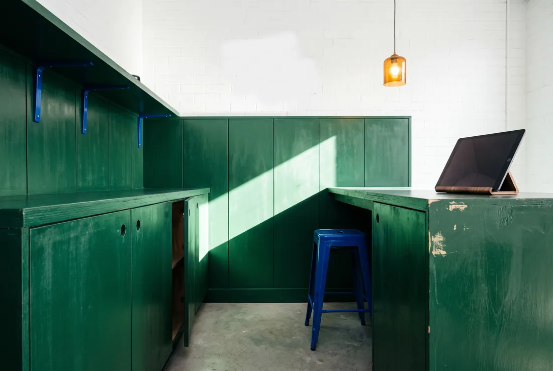 A corner of green painted joinery with a blue stool, an amber pendant lamp and a tablet on a stand.