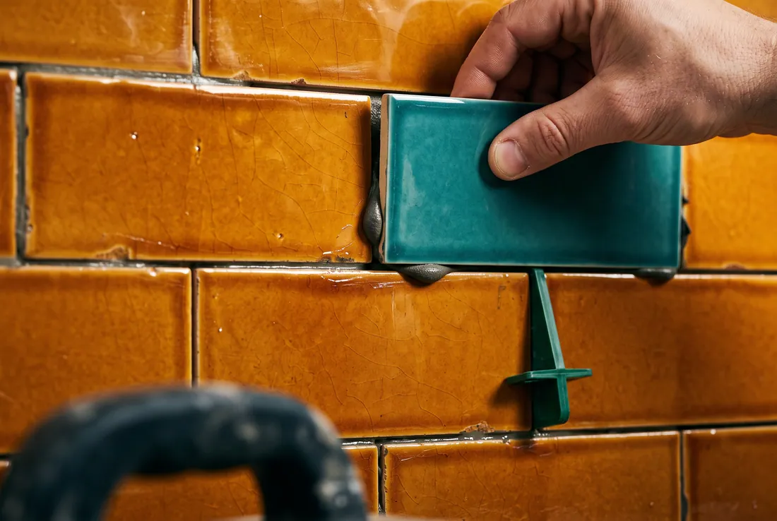 A hand setting a single teal glazed tile into a wall of amber tiles, a green spacer and adhesive at the joint.