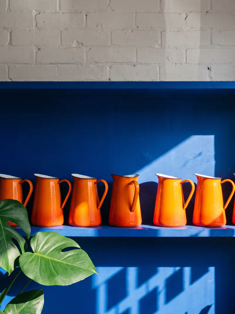 A row of glossy orange ceramic jugs lined up on a deep blue shelf beside a monstera leaf.