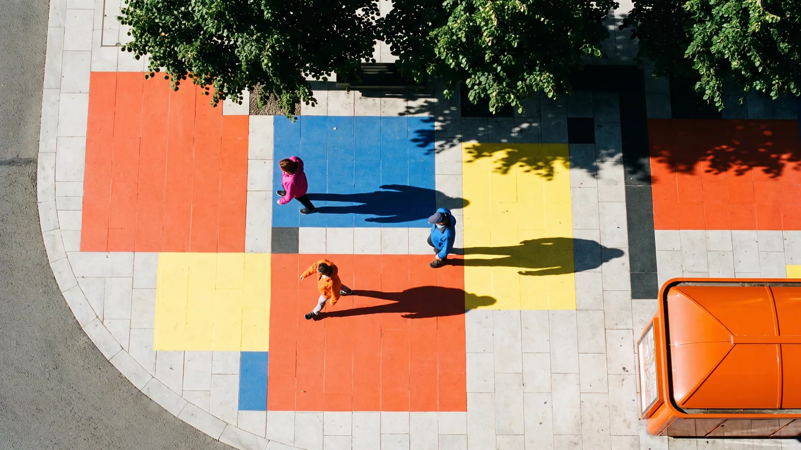 An aerial view of people walking across a plaza paved in orange, blue and yellow painted blocks under a tree.